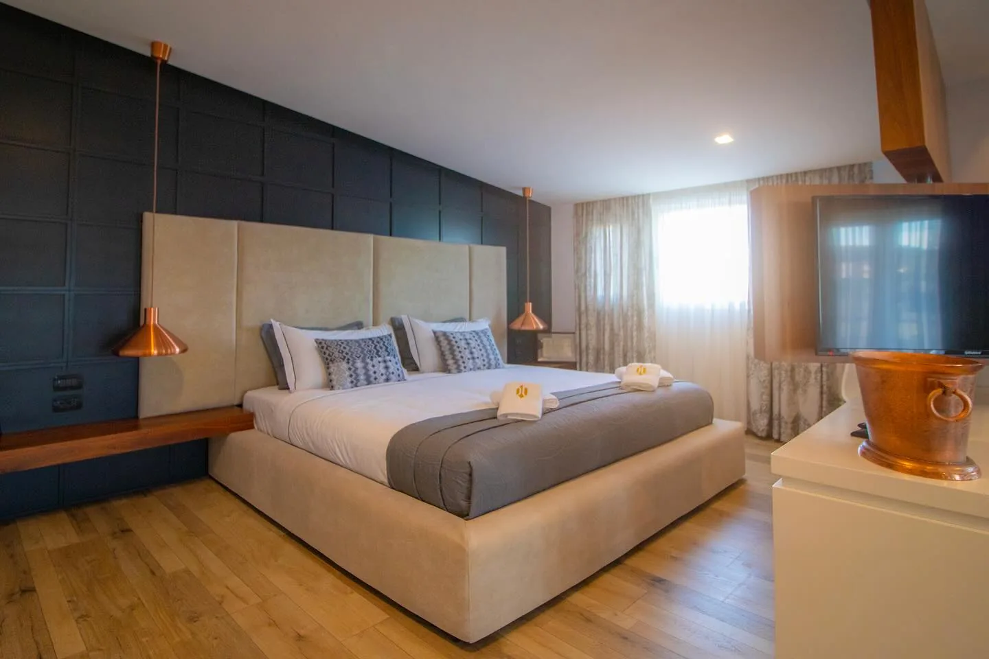 Bedroom with a cream padded headboard against a dark blue panelled wall, copper pendant lights and a wall-mounted television