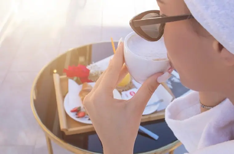 Person in robe and hair towel drinking cappuccino beside breakfast tray