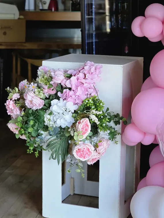Pink and white floral arrangement on white plinth beside balloons