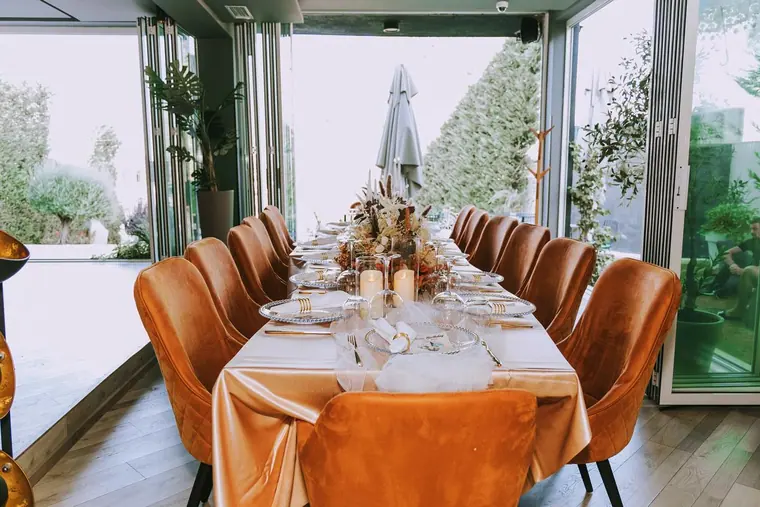Long banquet table with orange velvet chairs beside open glass doors to garden