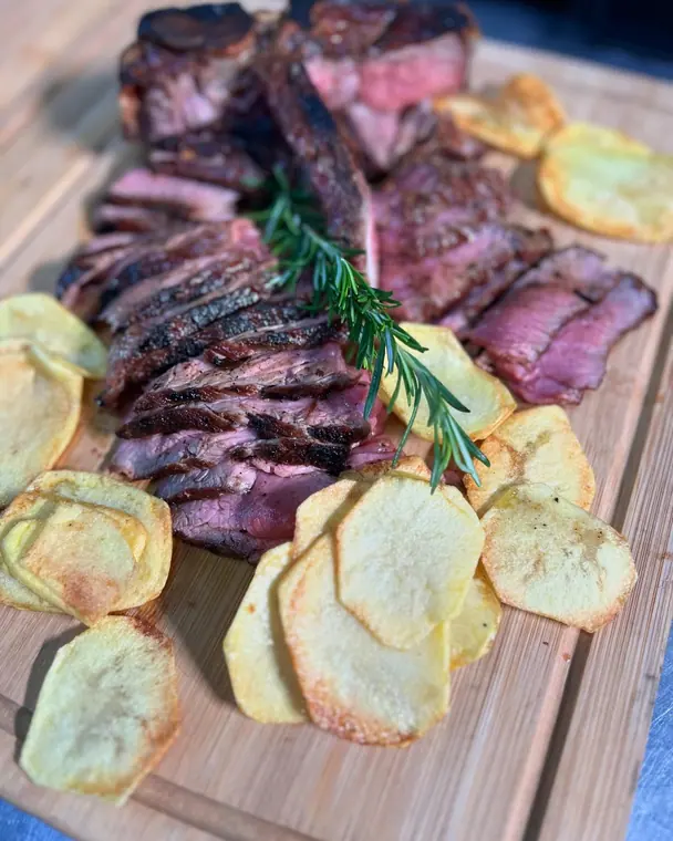 Sliced grilled steak with rosemary and potato crisps on a board