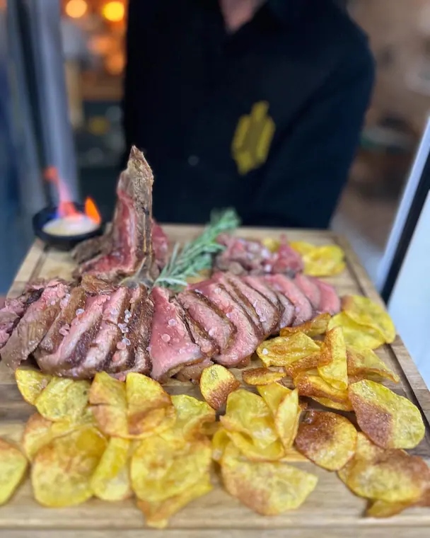Sliced steak on the bone with crisps on a board, flaming sauce pot