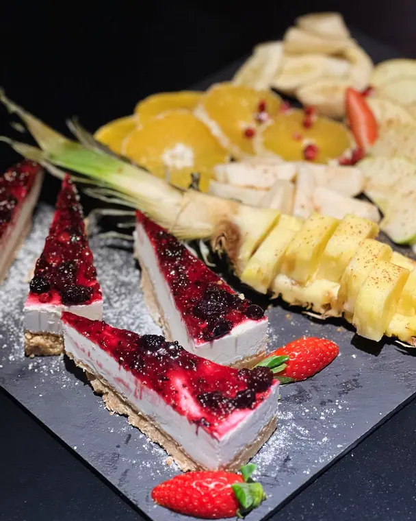 Three cheesecake slices with berries and fruit platter on slate